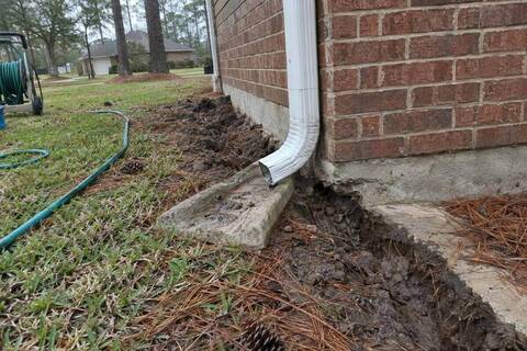 Downspout ending on a splash block beside a slab edge, dark clay soil and pine straw around it