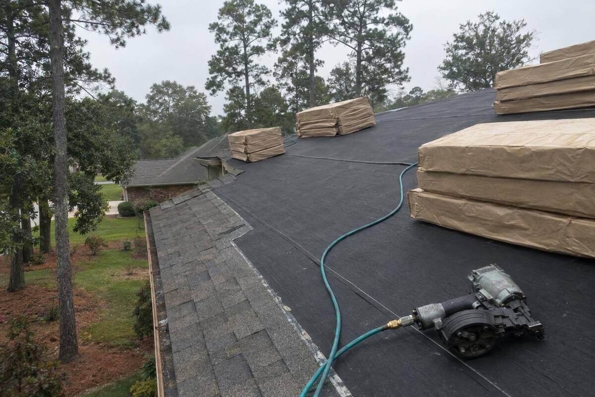 Bundles of laminated shingles stacked on a roof slope with tall pines behind